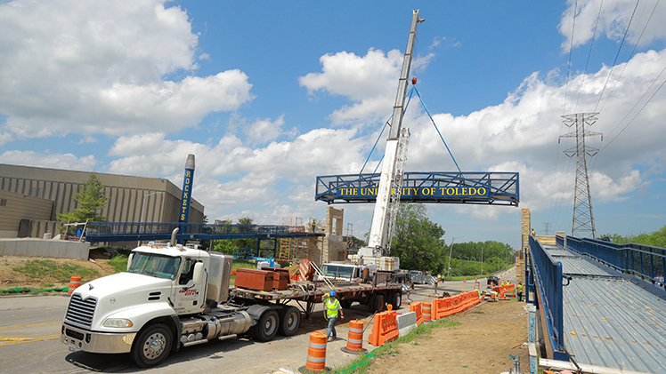 A white crane truck lifts the blue 78-foot-wide pedestrian bridge span into place over Douglas Road