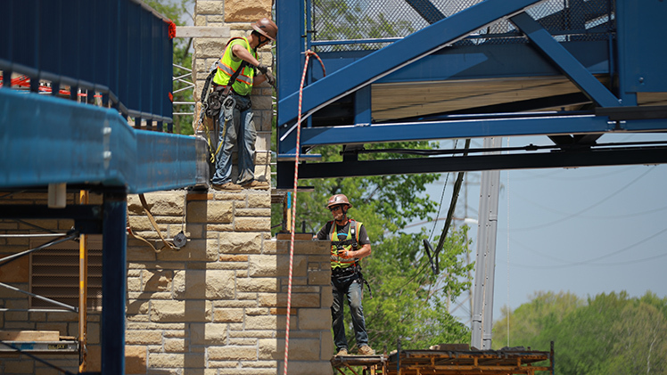 Construction workers wearing hardhats and reflective safety vests work to secure the bridge in place. 