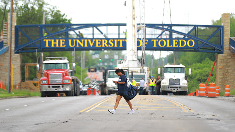 A pedestrian wearing UToledo gear crosses the closed section of Douglas Road in front of the new pedestrian bridge. 