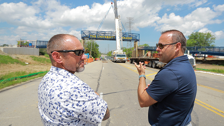 Pat Blevins, manager of structural maintenance for Main Campus, and Jason Toth, senior associate vice president for administration, stand on Douglas Road talking as a crane lifts pedestrian bridge span into place in front of them.