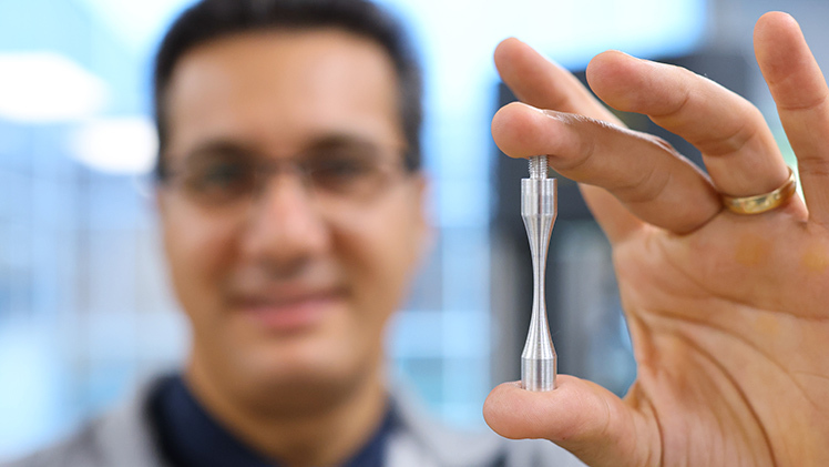 Dr. Meysam Haghshenas holds an hourglass test specimen for use in a fatigue tester inside his Failure, Fracture and Fatigue Laboratory.