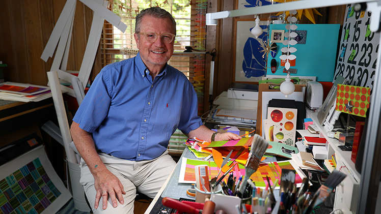 Portrait of local artist Steven Wipfli, who is posing at the desk in his home studio.