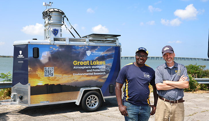 Dr. Kennedy Doro and Dr. David Kennedy stand in front of UToledo's trailer-mounted atmospheric sampling platform at the Lake Erie shoreline.