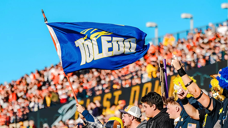 Photo of UToledo students at a football game with one student waving the Toledo Rockets flag.