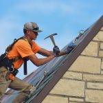 A roofing contractor wearing an orange shirt swings a hammer to nail down a new slate roof tile on MacKinnon Hall