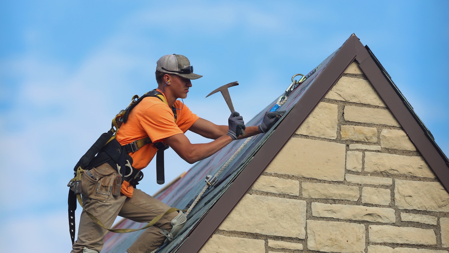 A roofing contractor wearing an orange shirt swings a hammer to nail down a new slate roof tile on MacKinnon Hall