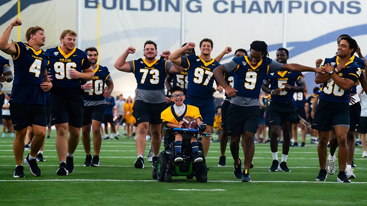 Rocket football players cheer on a child in a motorized wheelchair as he carries a football downfield inside the Fetterman Training Center.