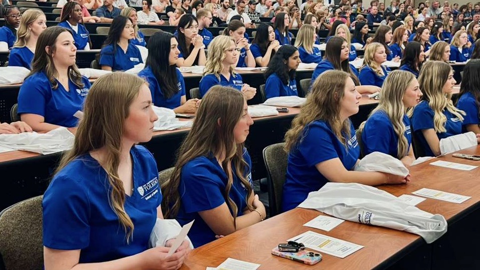 Nursing students wearing blue scrubs sit in an auditorium. 