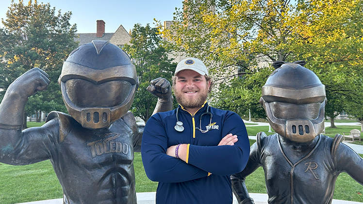 Senior nursing student Chase Gillson, wearing a stethoscope, poses in between the Rocky and Rocksy statutes.