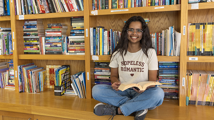 Portrait of Mariya Kottilangatt Jojy, a senior studying English with a concentration in literature, who is holding a book in the Carver Resource Center. She is using her internship at the Carver Resource Center to provide easy access to reading materials for young learners.