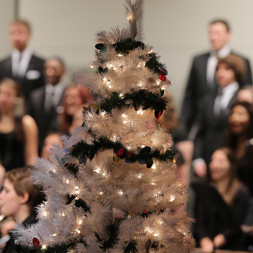 Close-up photo of a lit-up Christmas tree with singers in the background.