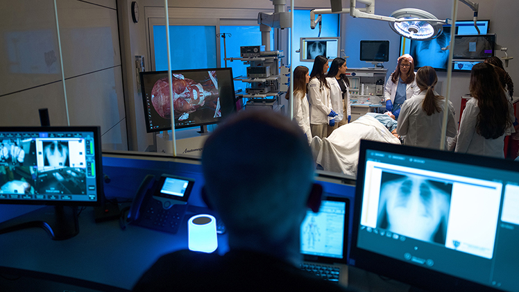 Instructor leads a group of medical students in white coats around a simulated patient in a high-tech operating room at The University of Toledo’s Interprofessional Immersive Simulation Center, as a technician monitors the session from a nearby control room with multiple screens showing medical imaging.