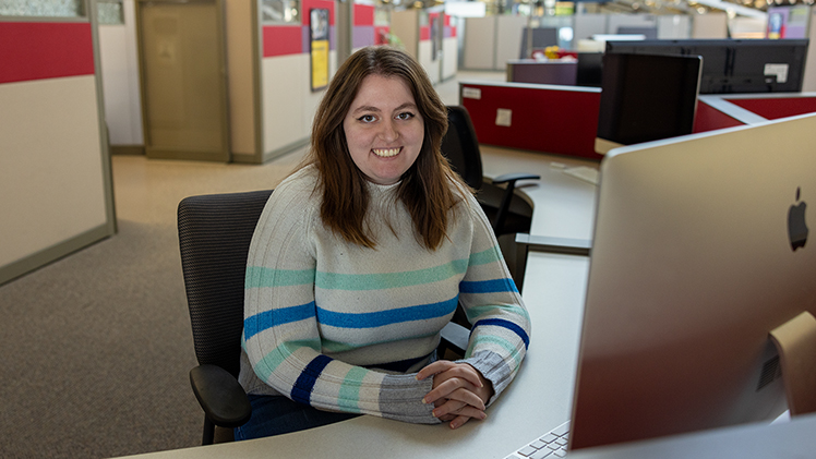 Portrait of Natalie Burgess, a student writer for University Marketing and Communications who graduates Dec. 13 with a bachelor’s degree in English, posing for a photo while sitting by a computer in the MARCOM office.