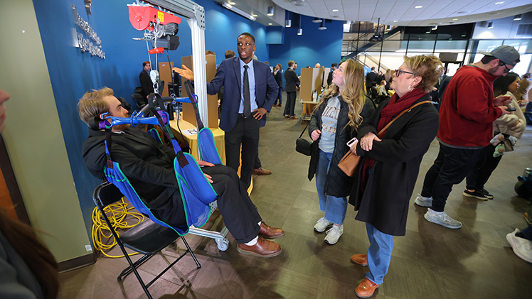 Ben Kasper, a mechanical engineering senior, demonstrates his team’s device to assist individuals who use wheelchairs into hotel beds as Ibraheem Oguntade, also a mechanical engineering senior, explains their project to Natalie Frankel and Elaine Stallard during the College of Engineering’s Senior Design Expo.