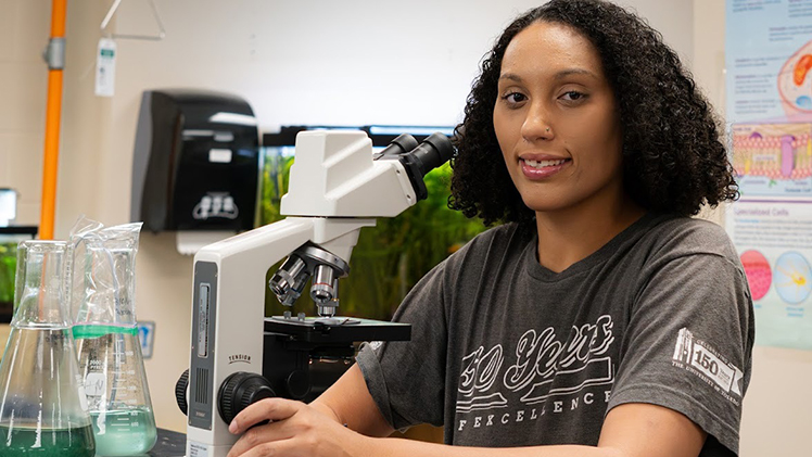 Photo of Tearica Moore at a desk with a microscope. She is graduating Saturday with a bachelor of science degree in biology.