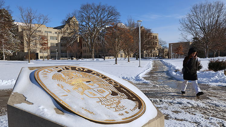 A student walks across a snowy Centennial Mall with the University's seal in the foreground covered with snow.