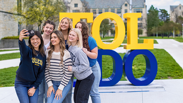Portrait of a group of female students posing for a selfie in front of the TOL-EDO sign on Main Campus.