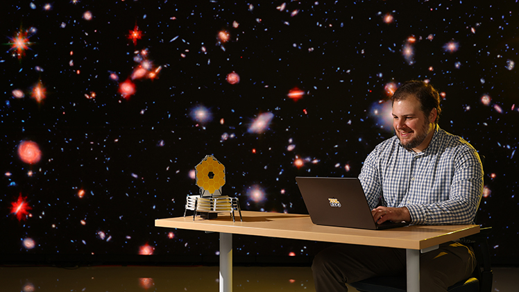 Dr. Eli Visbal, an associate professor and theoretical astronomer who led new research proposing the first direct detection of some of the oldest stars in the universe, sits at a computer with hundreds of images of spiral galaxies on the video wall behind him.