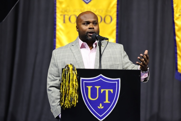 Bryan Blair, a man wearing a pink shirt and light gray blazer, stands behind a podium with the UToledo shield logo