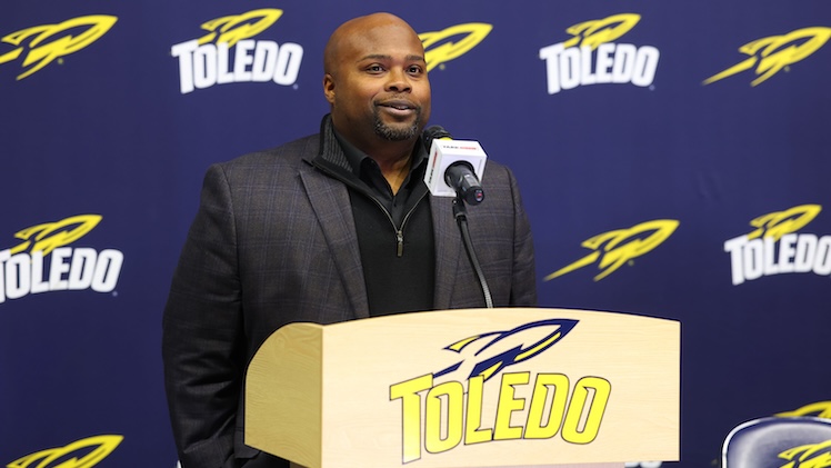 Bryan Blar, a man wearing a black shirt and gray blazer, stands behind a podium with the Toledo Rockets logo repeating on blue background behind him