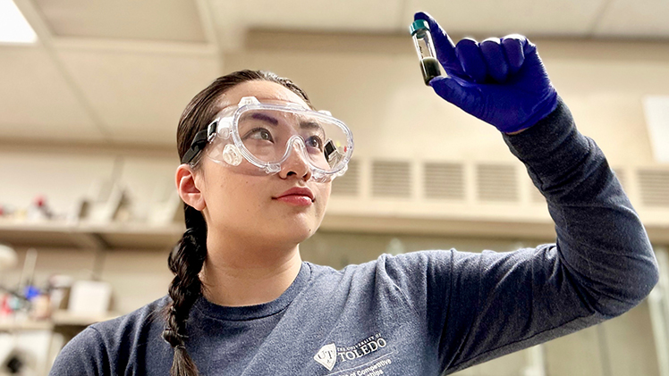 Photo of UToledo student Victoria Ho in a lab wearing goggles while looking at a small vial.