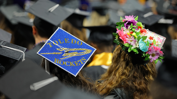 A photo of the tops of graduation caps at a UToledo commencement ceremony, with one cap saying "Toledo Rockets" with a rocket and another adorned with flowers.
