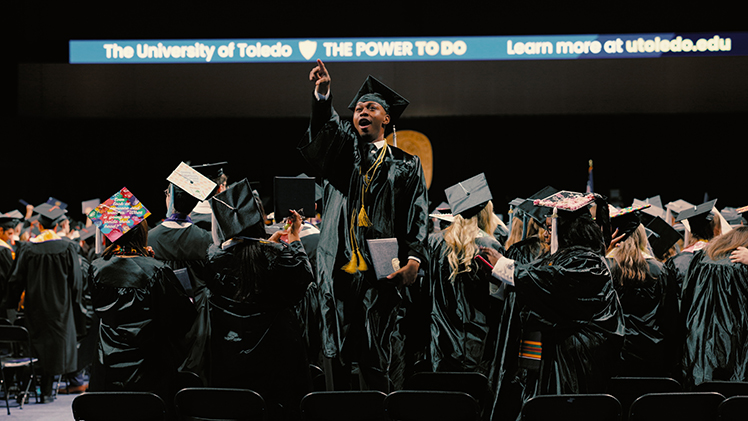 During a spring 2026 commencement ceremony, a UToledo student who has just graduated points to the stands in celebration.