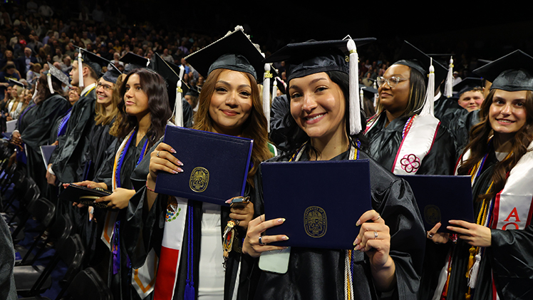 ALT: Two female UToledo graduates pose at a spring 2026 commencement ceremony  while holding their diploma placeholders.