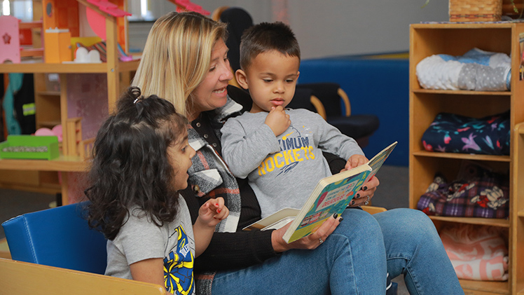 A female teacher in the UToledo Early Learning Center reads to a young boy and young girl.