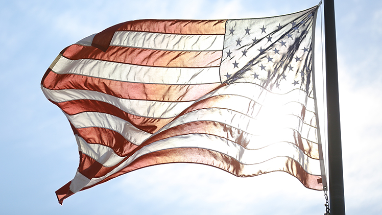Stock photo of the American Flag with the sun shining behind it. 