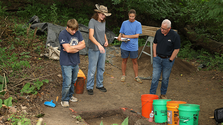 Undergraduates Hunter Hogan and Brayden Hickerson show the test unit they are excavating at a private-property site adjacent to Maumee’s Fort Maumee to Dr. Melissa Baltus, an associate professor of anthropology, and UToledo President James Holloway.