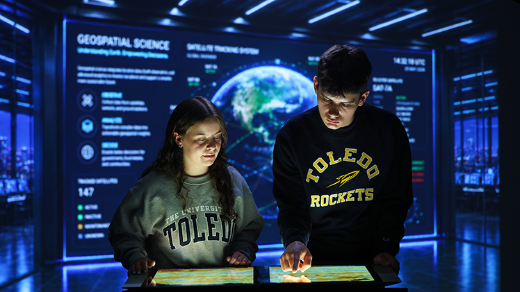 Two UToledo students look down at a glowing computer screen for information in a high-tech room setting.